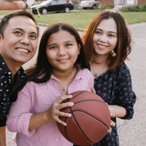 Young teen holding basketball is flanked by two adults. All are smiling into a camera for a selfie photo.